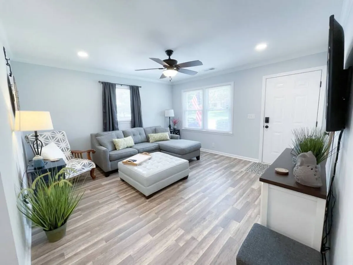 This image features a cozy living room with a plush sofa, an ottoman positioned for group gatherings, a window allowing natural light to fill the space, and a stylish lamp for ambient lighting. The set-up encourages relaxation, reading, and socializing.