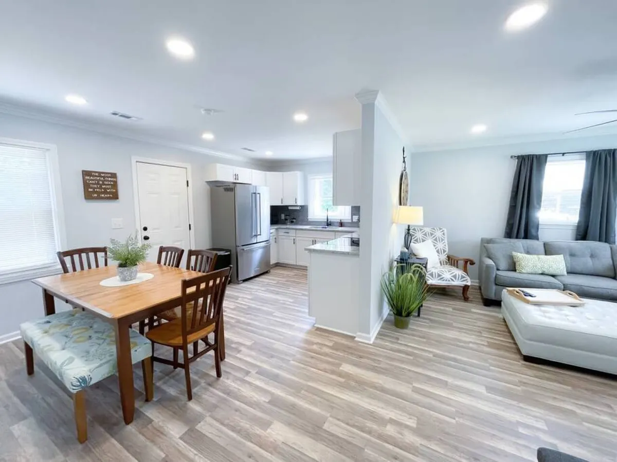 This image showcases a bright, open-plan living space with a dining table, kitchen area, and comfortable living room seating. A decorative plant adds a fresh touch, while natural light fills the room, creating a welcoming environment.