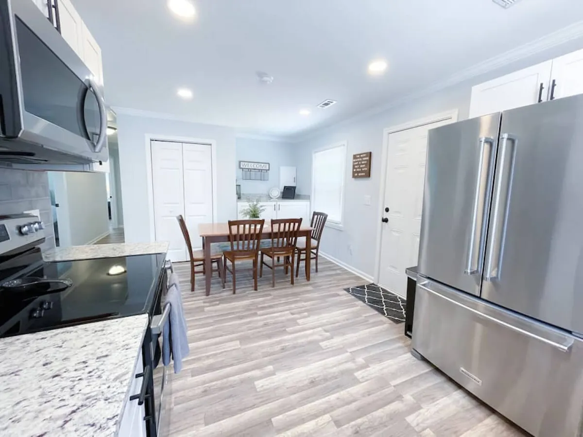 This image displays a dining area featuring a kitchen table with seating and decor, surrounded by kitchen appliances and bathed in natural light from nearby windows. The space feels open and ideal for group dining or casual meals.