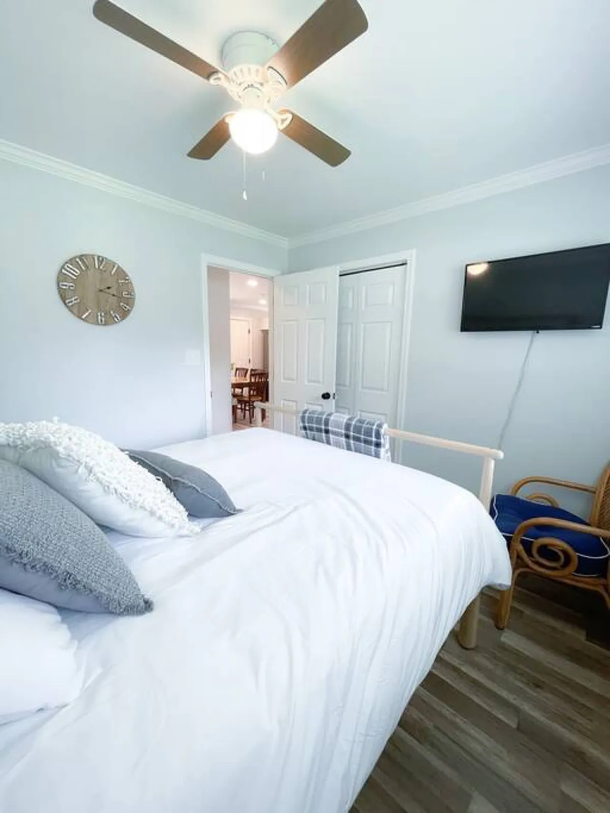 A cozy bedroom with decorative pillows, a wall clock, a TV mounted on the wall, and a comfortable chair in the corner. The natural light fills the room, enhancing the calm and inviting ambiance.