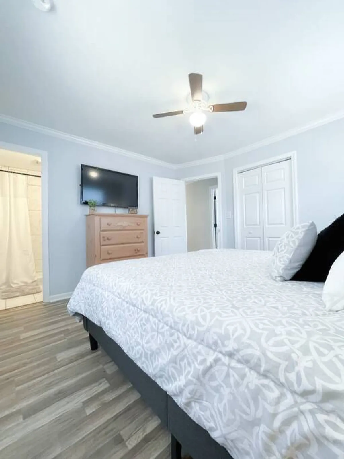 A serene bedroom with a comfortable bed dressed in decorative pillows, a ceiling fan overhead, and a dresser with a TV. The atmosphere is cozy and restful, ideal for winding down.