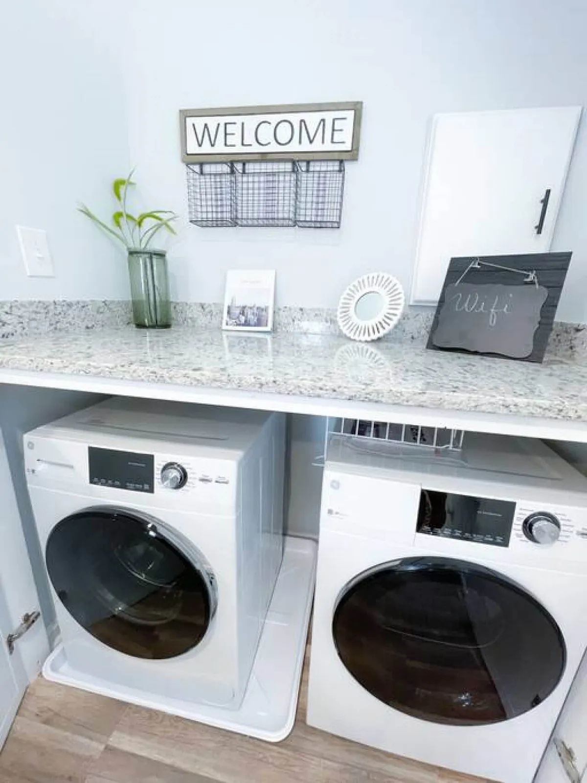 This image presents a functional laundry area complete with a washing machine, dryer, welcome sign, decorative plant, and a visible wifi sign. The organized, clean space hints at the amenities and thoughtful details provided for guests.