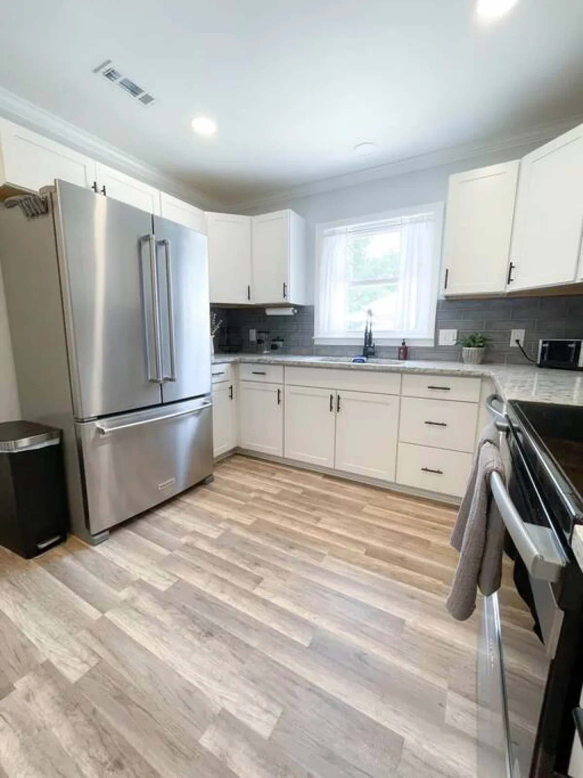 A bright, well-appointed kitchen with a stainless steel refrigerator, central island, and ample natural light streaming through the window. Open cabinets speak to convenience and modern living, and the space feels ready for entertaining or winding down after a busy day.