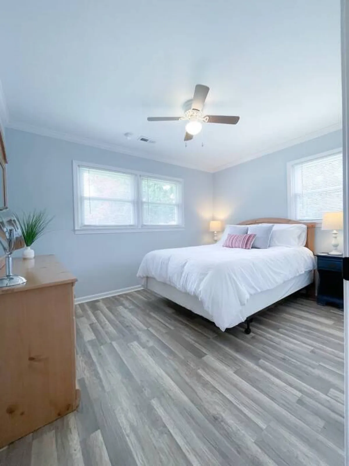A bright bedroom with white bedding, a wooden dresser, ceiling fan, and two sunlit windows. The overall atmosphere is restful and inviting, suitable for guests seeking a peaceful night's sleep.