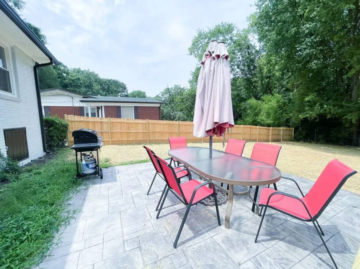 A vibrant backyard and patio scene with a dining table surrounded by red chairs, a large umbrella for shade, a propane grill ready for outdoor meals, and a fully fenced yard offering privacy and safety. The space is sunlit and welcoming, ideal for alfresco dining and relaxation.
