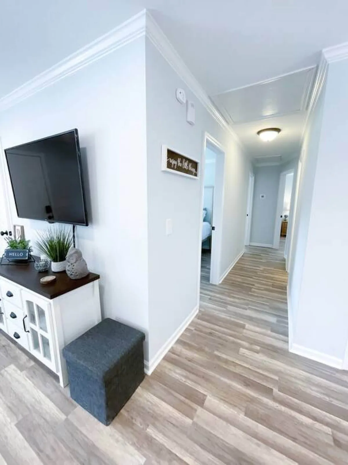 A welcoming living room featuring a large television, console table, decorative sign, and planters, with a hallway visible in the background. The arrangement invites relaxation and group gatherings.