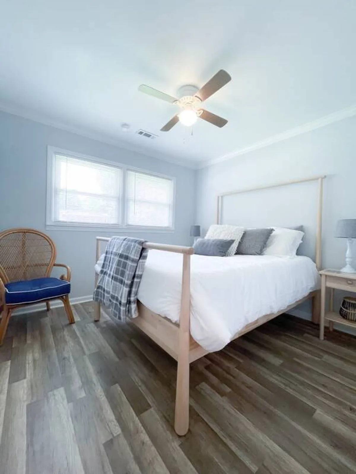 This image shows a tranquil bedroom featuring a comfortable bed with crisp white bedding, a blue accent chair, wooden floors, a ceiling fan, and ample natural light coming through the window. The scene evokes a sense of peace and relaxation, highlighting the restful spaces available in the home.